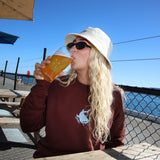 Person drinking a beer at an outdoor setting with a clear blue sky and a seagull.