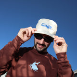 Man wearing a brown sweatshirt with a logo and a cream bucket hat with 'Gage' text against a blue sky.